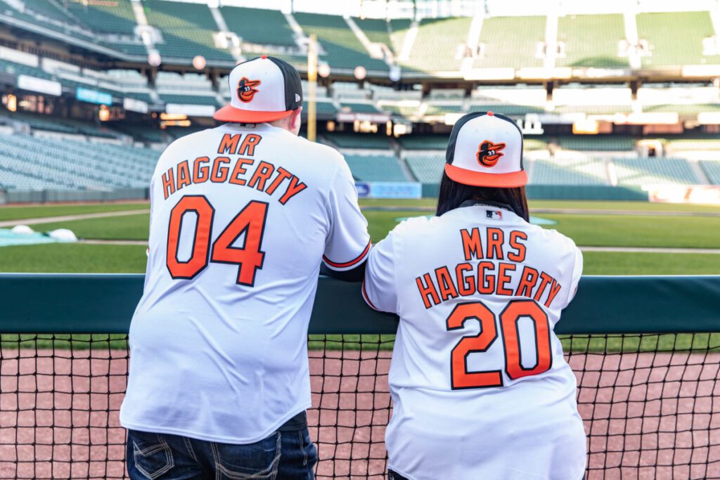 Camden-Yards-Engagment-34 - TOMMY GATZ ENTERTAINMENT Two people in matching baseball jerseys stand at a stadium's edge, looking at the field. Jerseys read "Mr. Haggerty 04" and "Mrs. Haggerty 20.
