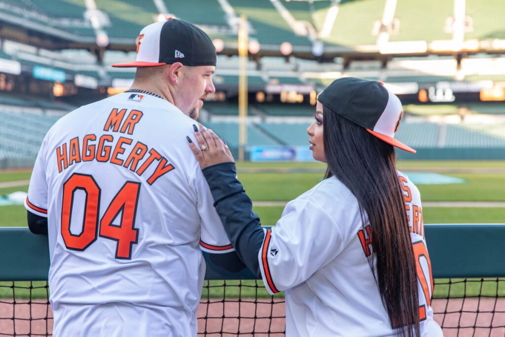 Camden-Yards-Engagment-35 - TOMMY GATZ ENTERTAINMENT A couple in matching baseball jerseys, with "Mr. Haggerty" and "Mrs. Haggerty" on the back, stands in a stadium facing the field.
