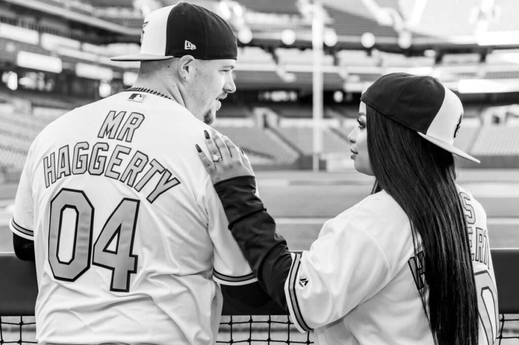 Camden-Yards-Engagment-36 - TOMMY GATZ ENTERTAINMENT A couple in matching baseball jerseys stand at a sports stadium, with the back of their shirts reading "Mr. Haggerty 04" and "Mrs. Haggerty." They are looking at each other.