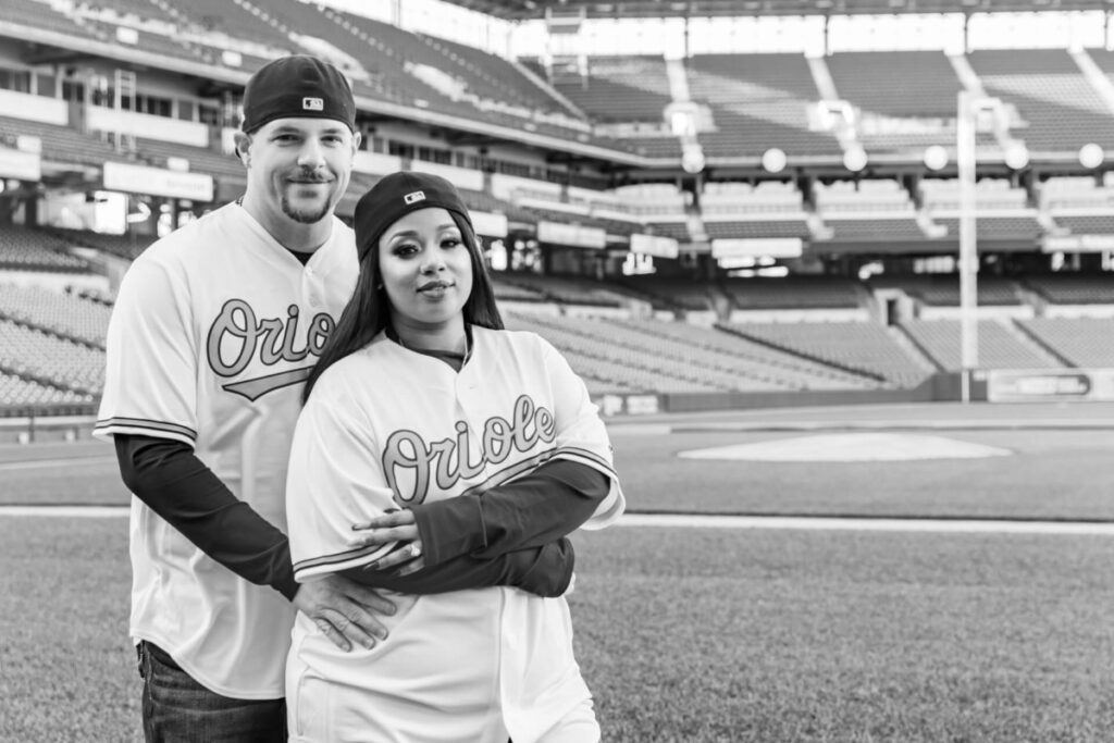 Camden-Yards-Engagment-39 - TOMMY GATZ ENTERTAINMENT A man and woman in Orioles jerseys pose together on a baseball field with empty stands in the background.