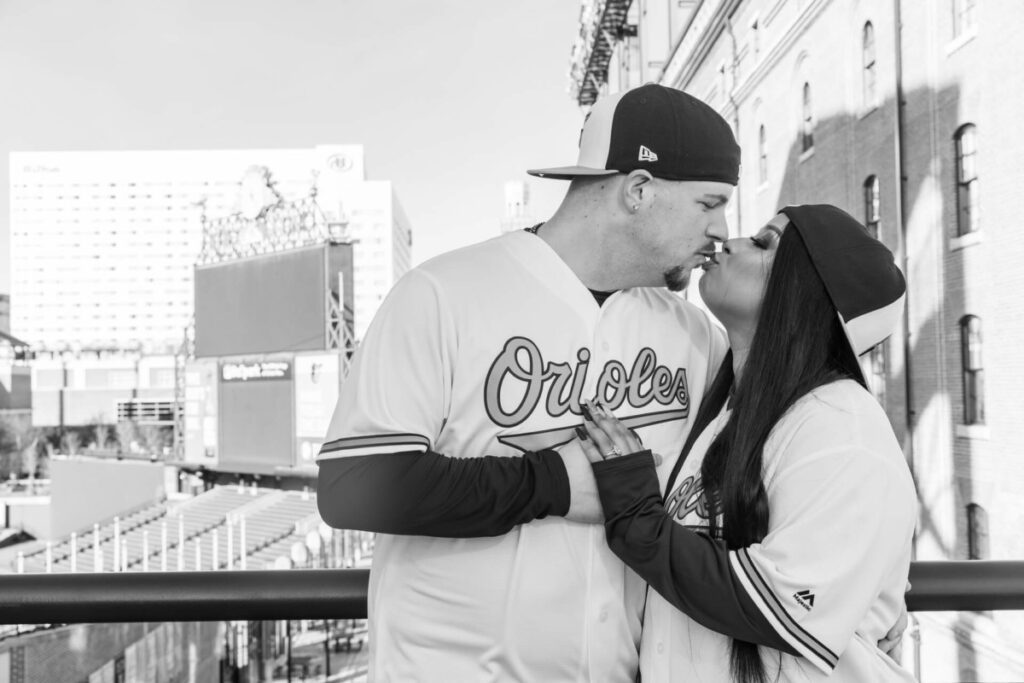 Camden-Yards-Engagment-4 - TOMMY GATZ ENTERTAINMENT A couple wearing Orioles jerseys kisses on a balcony outside a stadium.