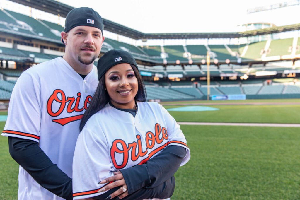 Camden-Yards-Engagment-40 - TOMMY GATZ ENTERTAINMENT A couple in Orioles baseball jerseys stands on a baseball field. The man is embracing the woman from behind as they both smile at the camera.