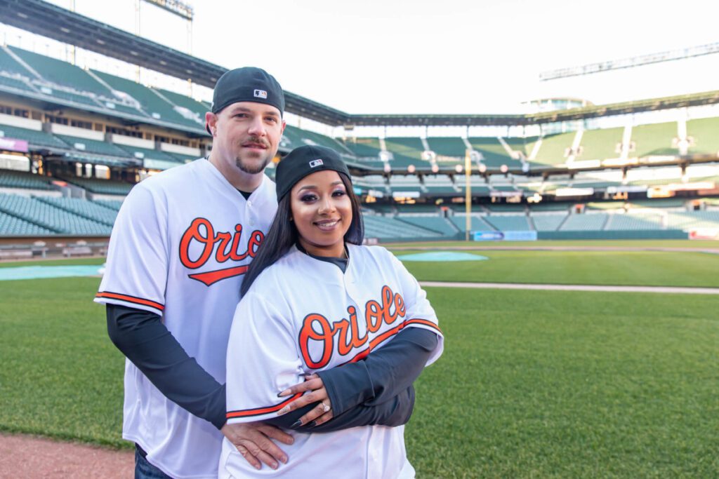 Camden-Yards-Engagment-41 - TOMMY GATZ ENTERTAINMENT Two people in "Orioles" baseball jerseys stand on a green field in an empty stadium. The person in front wears a black cap.