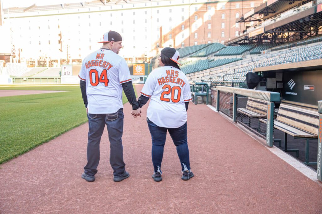 Camden-Yards-Engagment-42 - TOMMY GATZ ENTERTAINMENT A couple in matching jerseys labeled "Mr. Haggerty 04" and "Mrs. Haggerty 20" holds hands, walking through a baseball stadium.