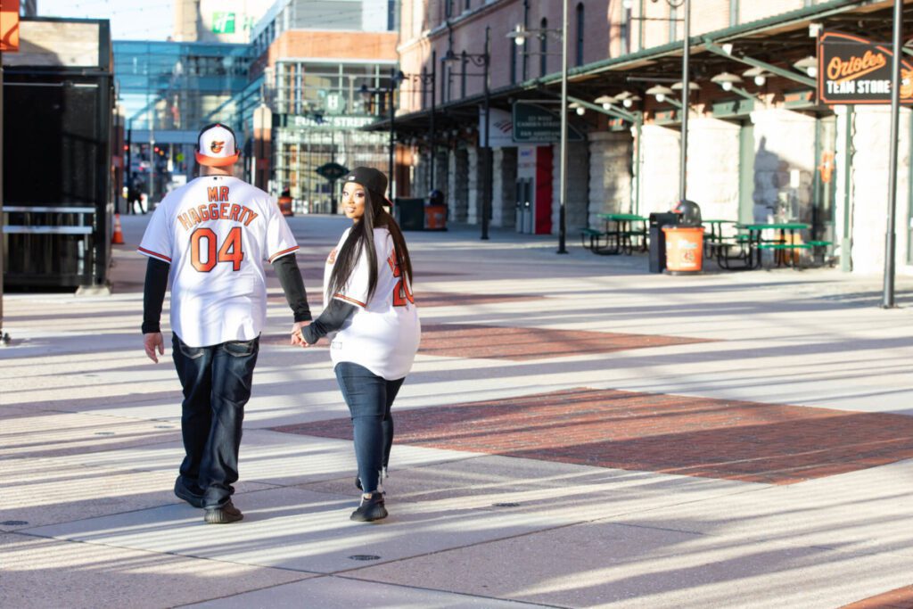 Camden-Yards-Engagment-44 - TOMMY GATZ ENTERTAINMENT A couple wearing matching baseball jerseys walks hand-in-hand along a sunlit street near a team store.