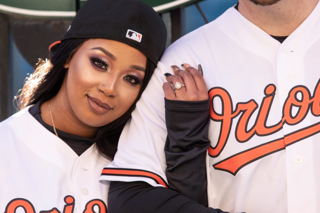 Camden-Yards-Engagment-46 - TOMMY GATZ ENTERTAINMENT A person in an Orioles baseball jersey and cap leans against another person wearing a matching jersey, showing an engagement ring on their left hand.