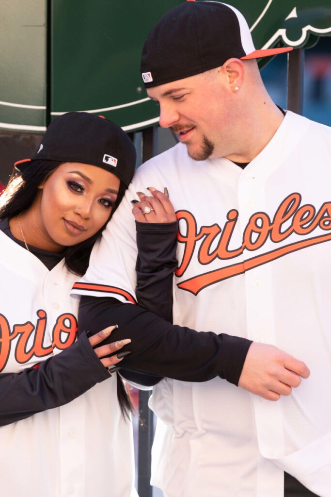Camden-Yards-Engagment-47 - TOMMY GATZ ENTERTAINMENT Two people wearing Orioles jerseys and caps pose together, with the woman leaning on the man's shoulder.