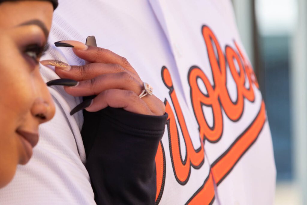 Camden-Yards-Engagment-48 - TOMMY GATZ ENTERTAINMENT Close-up of a person resting their hand on someone wearing an Orioles jersey. A ring is visible on the person's finger.