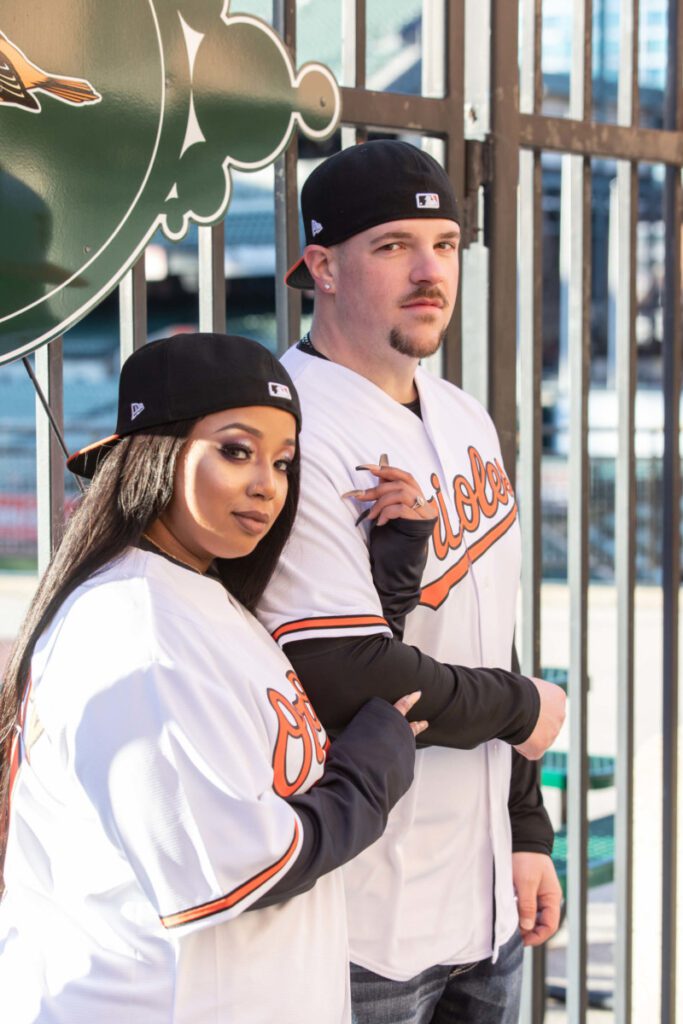 Camden-Yards-Engagment-49 - TOMMY GATZ ENTERTAINMENT Two people in baseball jerseys and caps stand in front of a metal gate, with one person crossing their arms and the other resting a hand on the gate.
