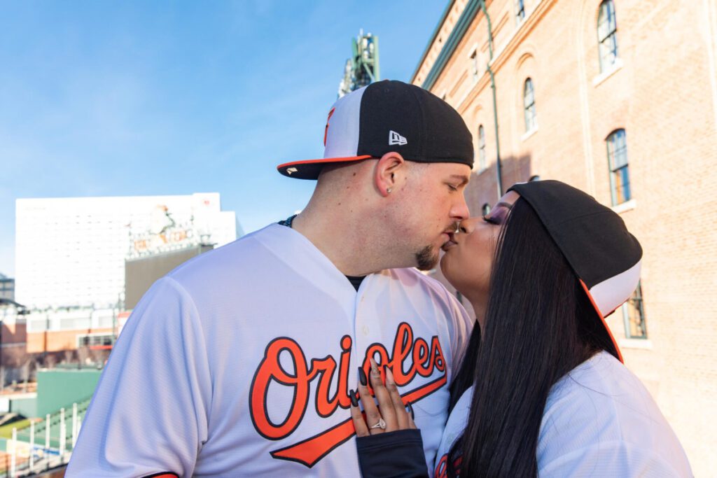 Camden-Yards-Engagment-5 - TOMMY GATZ ENTERTAINMENT A couple in Orioles jerseys kisses outside a baseball stadium.