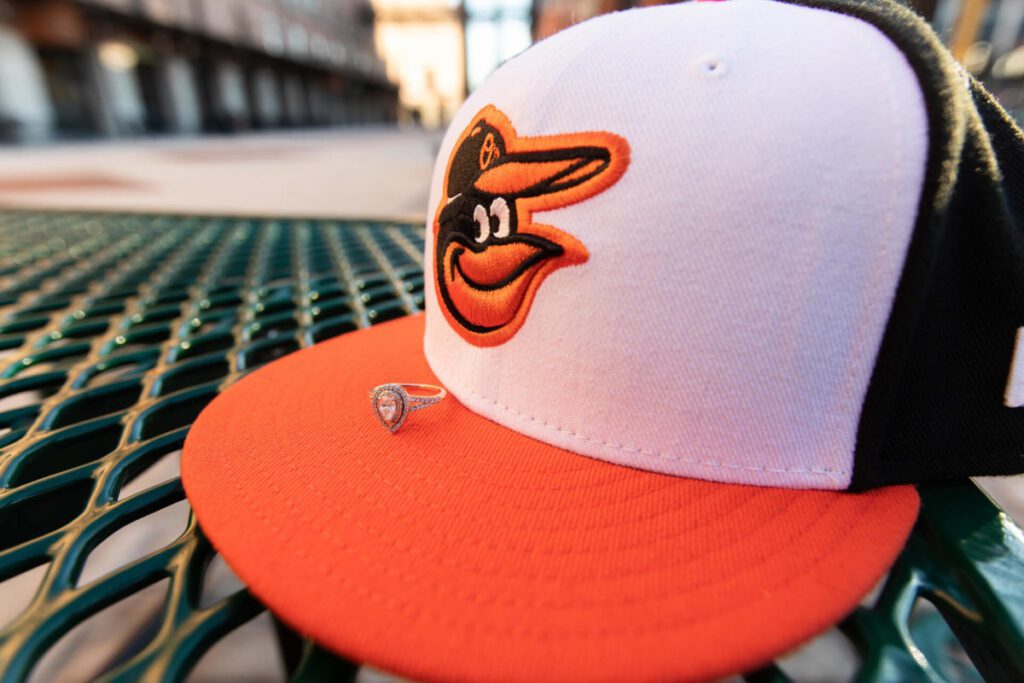 Camden-Yards-Engagment-50 - TOMMY GATZ ENTERTAINMENT A baseball cap with an orange and black bird logo is on a green metal table. A diamond ring is placed on the cap's brim.