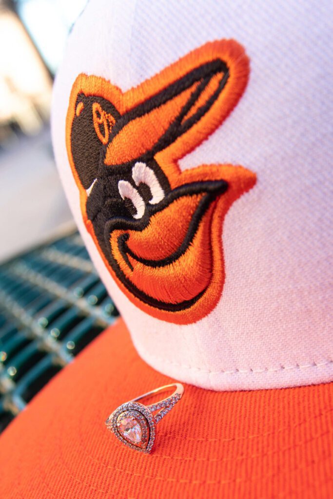 Camden-Yards-Engagment-51 - TOMMY GATZ ENTERTAINMENT White and orange baseball cap with an embroidered bird logo, placed on a metal bench. A diamond ring rests on the cap's brim.