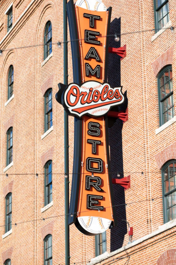 Camden-Yards-Engagment-52 - TOMMY GATZ ENTERTAINMENT Vertical "Orioles Team Store" sign on a brick building wall, featuring baseball imagery.