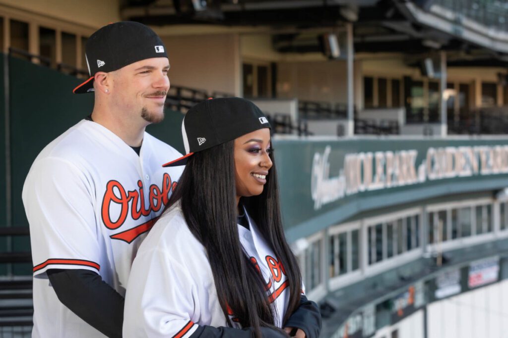Camden-Yards-Engagment-6 - TOMMY GATZ ENTERTAINMENT A man and woman in Orioles jerseys and caps pose together in a baseball stadium.