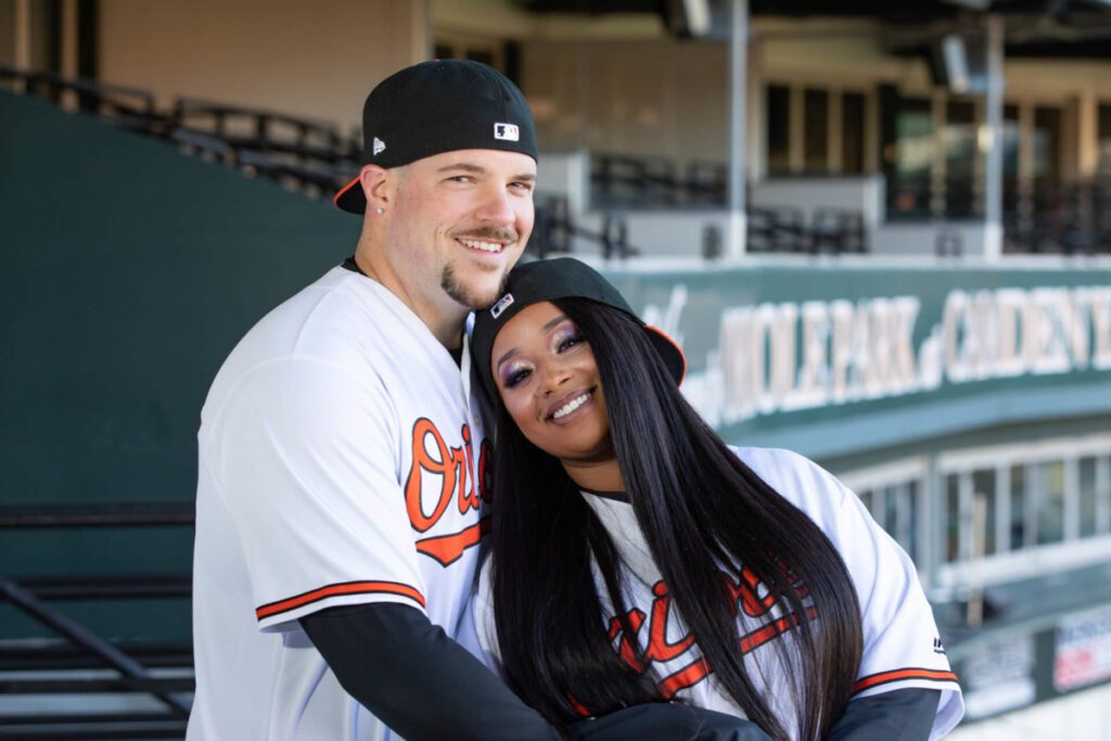 Camden-Yards-Engagment-7 - TOMMY GATZ ENTERTAINMENT Two people in Orioles shirts pose together at a baseball stadium. Both wear baseball caps; one is backward.