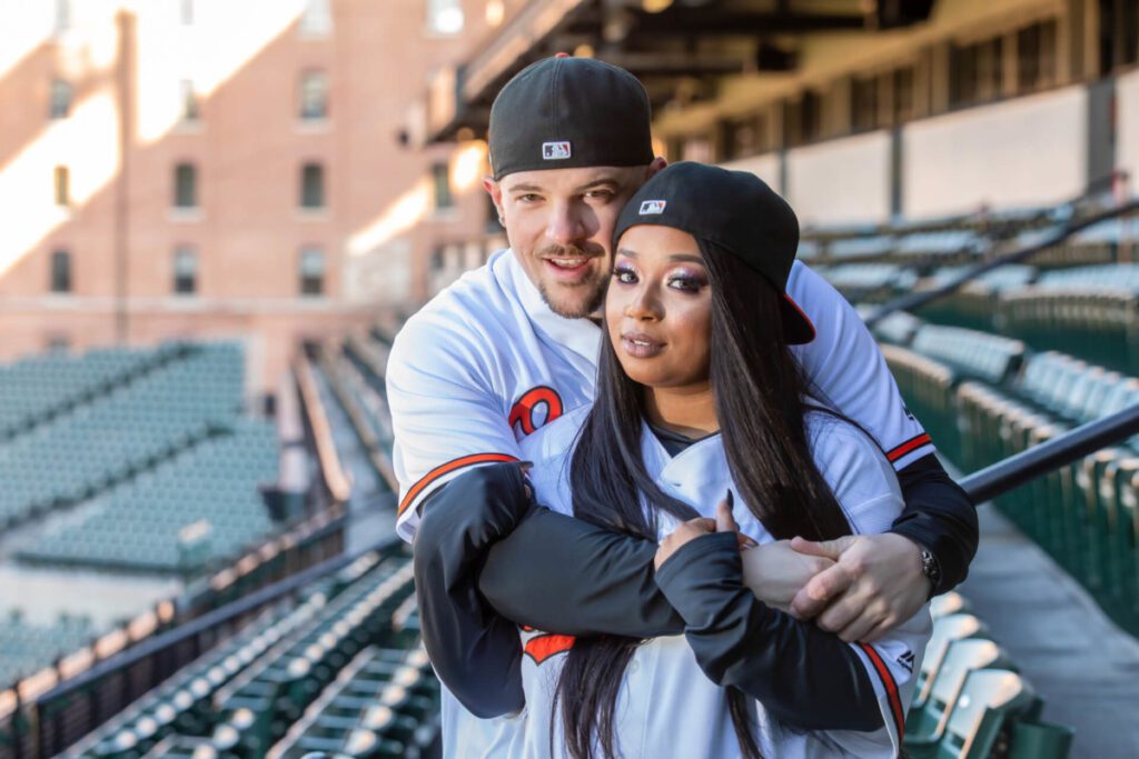 Camden-Yards-Engagment-8 - TOMMY GATZ ENTERTAINMENT A couple wearing baseball jerseys and caps are embracing in an empty stadium.