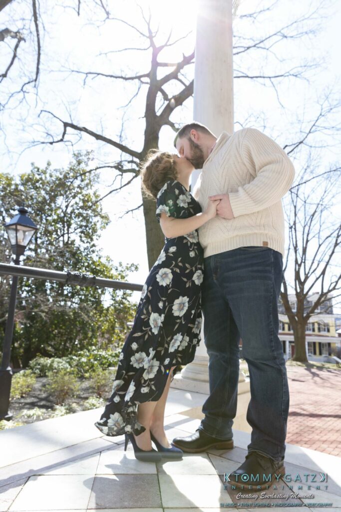 Shannon-James-031319-FINALS-Low-Res-WM-11 - TOMMY GATZ ENTERTAINMENT A couple kisses under a tall white column with bare trees and a lamp post in the background. The woman wears a floral dress; the man wears a white sweater and jeans.