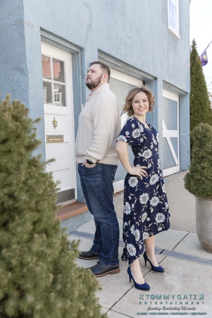 Shannon-James-031319-FINALS-Low-Res-WM-2 - TOMMY GATZ ENTERTAINMENT A man and a woman stand back-to-back outdoors. He wears a sweater and jeans; she wears a floral dress and heels. A blue building and greenery are in the background.