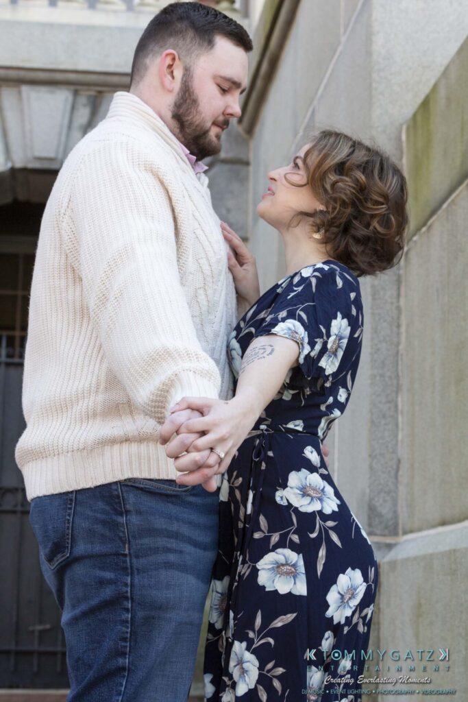 Shannon-James-031319-FINALS-Low-Res-WM-25 - TOMMY GATZ ENTERTAINMENT A couple holding hands and gazing at each other stands near a stone building. The man wears a cream sweater and jeans, and the woman wears a navy floral dress.