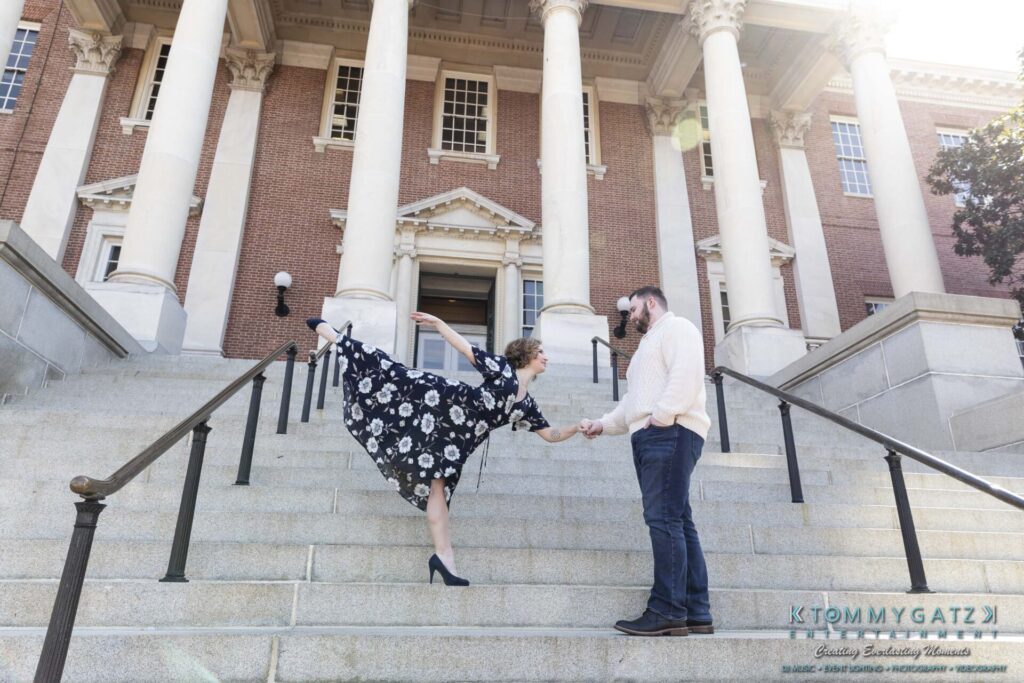 Shannon-James-031319-FINALS-Low-Res-WM-26 - TOMMY GATZ ENTERTAINMENT A woman in a floral dress playfully balances on one leg, holding hands with a man on the steps of a large brick building with tall columns.