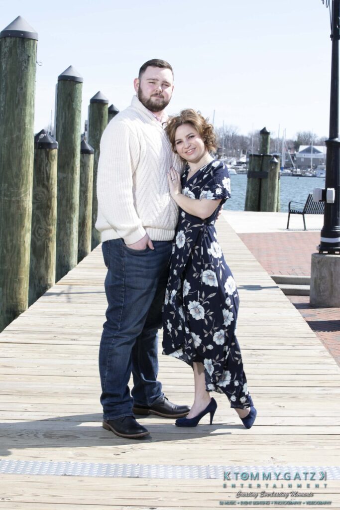 Shannon-James-031319-FINALS-Low-Res-WM-27 - TOMMY GATZ ENTERTAINMENT A couple stands on a wooden dock by the water, with the woman in a floral dress and the man in a white sweater and jeans. The background features posts and a streetlamp.