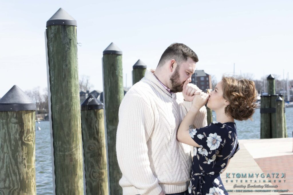 Shannon-James-031319-FINALS-Low-Res-WM-28 - TOMMY GATZ ENTERTAINMENT A couple stands by a waterfront, holding hands and leaning close. The man wears a cream sweater and the woman wears a floral dress. Dock posts and water are in the background.