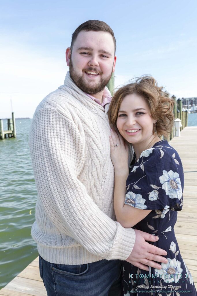 Shannon-James-031319-FINALS-Low-Res-WM-29 - TOMMY GATZ ENTERTAINMENT A couple poses on a wooden dock by the water; the man wears a cream sweater, and the woman wears a floral dress. They are smiling and embracing with the lake and piers in the background.