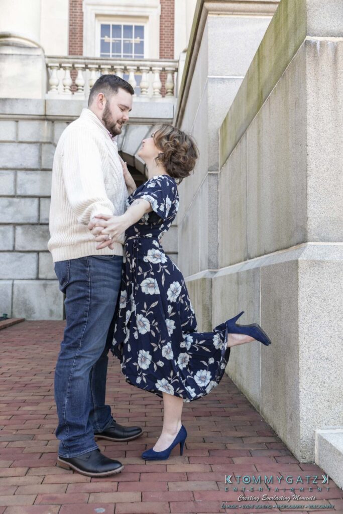 Shannon-James-031319-FINALS-Low-Res-WM-9 - TOMMY GATZ ENTERTAINMENT A couple poses affectionately near a brick wall. The woman in a blue floral dress holds the man's hands and lifts one leg. He wears a cream sweater and jeans.