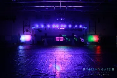 Dimly lit indoor venue with colorful stage lights in purple, blue, and red casting reflections on the floor, creating a nightclub atmosphere.