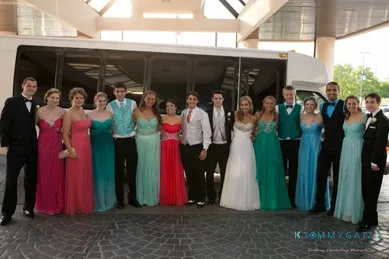 A group of young people in formal attire stand in front of a bus, posing for a photo. The girls wear colorful dresses and the boys are in suits and tuxedos.