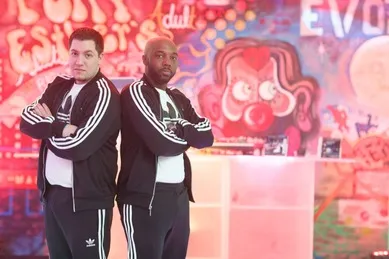 Two men in matching black and white tracksuits stand back-to-back with arms crossed. The background features colorful graffiti art.