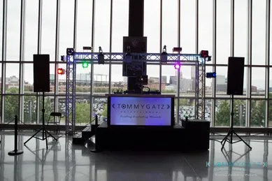 Stage setup with speakers, lights, and a DJ booth displaying "Tommy Gatz." Large windows in the background reveal an urban skyline.