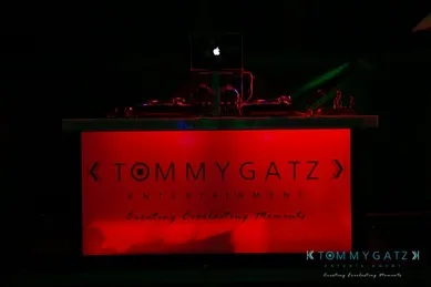 A DJ booth with a glowing red panel displaying "TOMMY GATZ ENTERTAINMENT" and an Apple laptop on top.