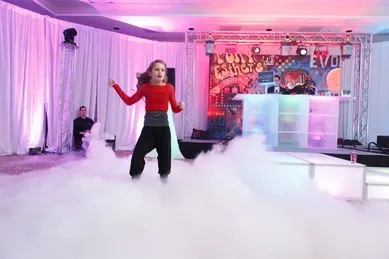 A person dances energetically on a stage surrounded by fog, with colorful lights and graffiti-style backdrop in the background.