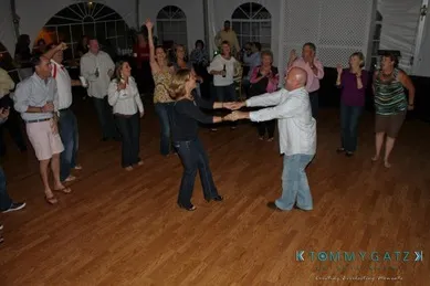 Group of people dancing energetically in a dimly lit room with wooden floors and arched windows.