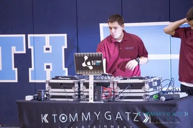 A DJ in a maroon shirt is adjusting equipment on a table with audio gear and a laptop. A person stands to the right, partially out of frame.