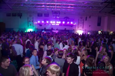 A large crowd of people dancing and mingling at an indoor event with purple stage lighting in the background.