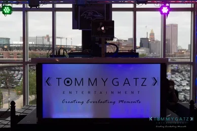 A DJ booth with "Tommy Gatz Entertainment" signage, set against a cityscape backdrop with cloudy skies.