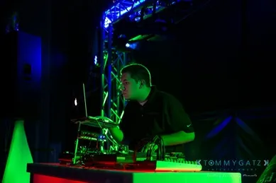 DJ performing on stage, surrounded by vibrant green and blue lights, with headphones on, using a laptop and mixer.