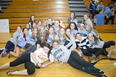 A group of young people in casual clothing pose together on a gym floor in front of wooden bleachers.