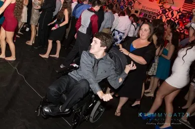 A wheelchair user in a gray shirt dancing at a crowded event with people in formal attire.