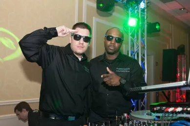 Two men wearing sunglasses pose near DJ equipment at an indoor event. One man salutes with his hand, and the other points towards the camera.
