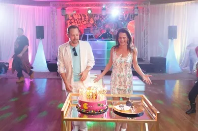 A man and woman stand behind a lit birthday cake on a table at a decorated indoor party.