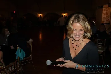 A woman holding a microphone stands in a dimly lit room, smiling. She is wearing a black dress and necklace.
