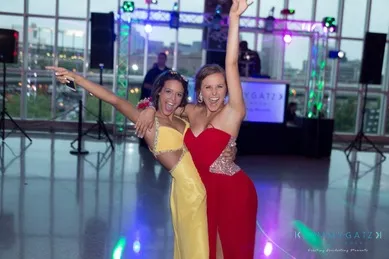 Two people in formal dresses pose with arms raised, smiling. The venue has glass walls and colorful lights in the background.