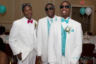 Three men in white suits with turquoise bow ties, two wearing sunglasses, pose together in a decorated event setting with balloons in the background.