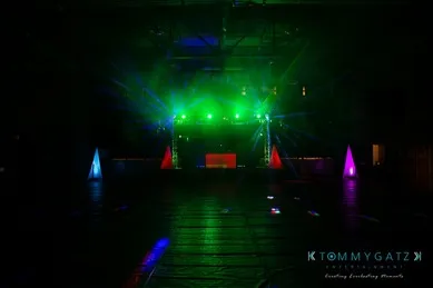 A dark event space with green and purple stage lighting, featuring triangular light structures and a digital screen.