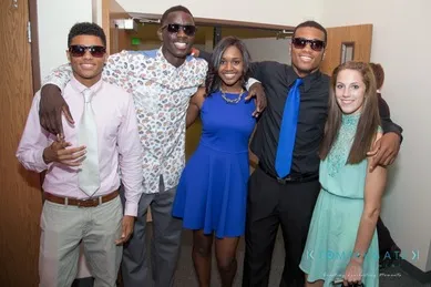 Five people standing together, smiling, dressed in semi-formal clothing. Four are wearing sunglasses, and two of the individuals are wearing blue outfits.