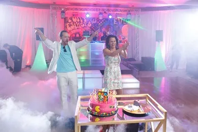 A man and woman celebrate in a party room with fog and colorful lights. A decorated cake sits on a table in the foreground.
