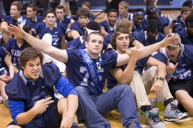 A group of young men in football jerseys sit on a gym floor, with one wearing a "Homecoming King" sash, posing with their arms raised.